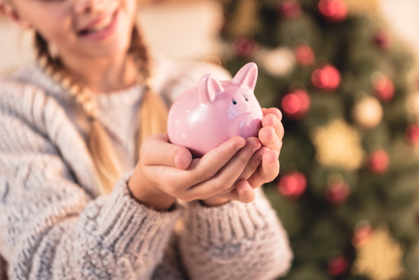 cropped view of kid holding piggy bank at home with christmas tree - Foto, Imagem