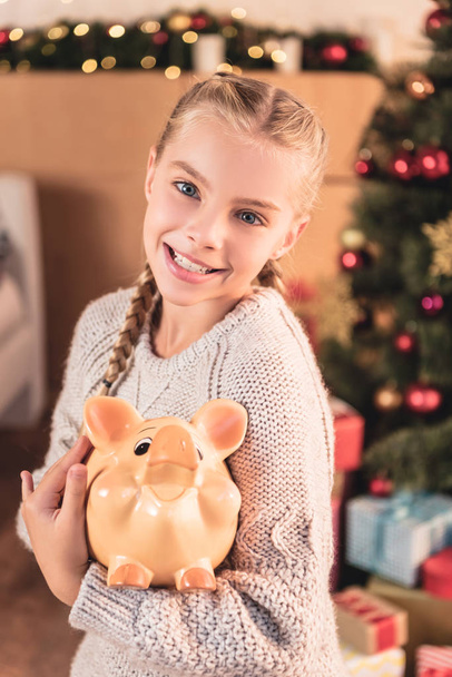 adorable smiling kid holding piggy bank at home with christmas tree - Fotoğraf, Görsel