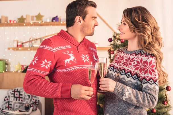 lachende paar in Kerstmis Truien champagneglazen houden en kijken naar elkaar - Foto, afbeelding
