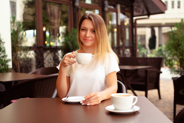 Portrait of the young beautiful woman looking at the camera and drinking a cup of cappuccino at the table on the summer veranda in a cafe. Pretty woman with cup of coffee. - Foto, imagen