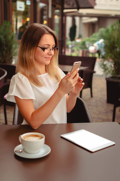 The young beautiful woman holding her mobile phone and reading or typing on it. She is sitting in the cafe with the cup of cappucino and digital tablet laying on the table. - Foto, Bild