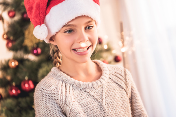 smiling preteen kid in santa hat holding sparkler at home with christmas tree - Fotó, kép