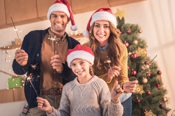 smiling parents and daughter in santa hats holding sparklers at home with christmas tree - Zdjęcie, obraz