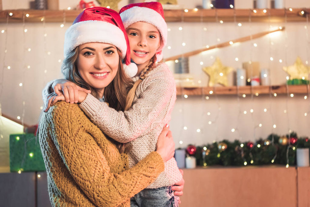 усміхнена мати і дочка в капелюхах Санти обіймаються вдома в різдвяний час
 - Фото, зображення