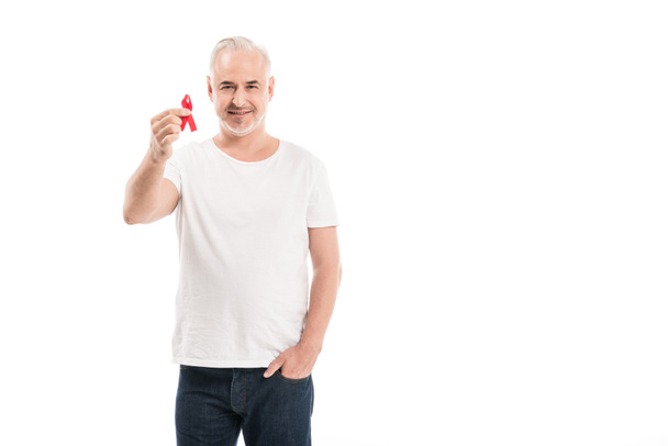 χαμογελώντας ώριμος άνδρας σε κενό λευκό t-shirt εκμετάλλευση του aids ευαισθητοποίησης κόκκινη κορδέλα και κοιτάζοντας κάμερα απομονωθεί σε λευκό - Φωτογραφία, εικόνα