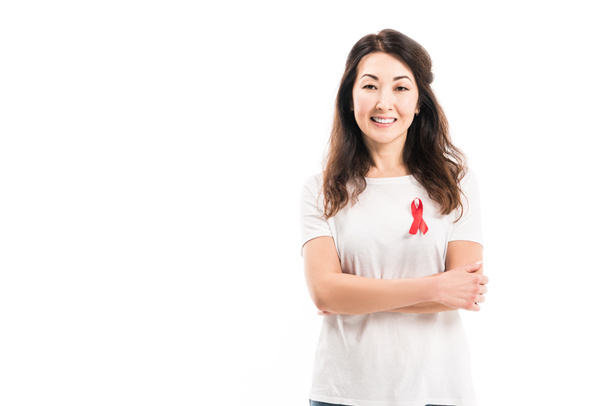smiling adult asian woman with aids awareness red ribbon on t-shirt looking at camera with crossed arms isolated on white - Photo, Image