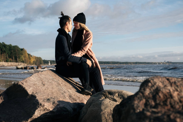 Couple in love sitting on the stone near the ocean, hugging and enjoy each other. Coastline on the background. Young man wearing a wool sweater, girl in a pink coat - Photo, Image