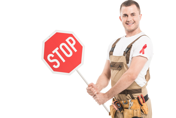 happy adult builder with aids awareness red ribbon on overall holding stop road sign and looking at camera isolated on white - Photo, Image