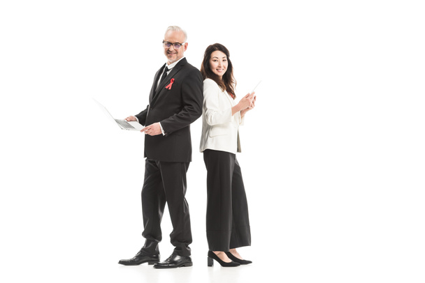 successful adult businesspeople with aids awareness red ribbons working with gadgets while standing back to back and looking at camera isolated on white - Photo, Image