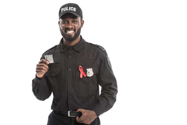 smiling african american police officer with aids awareness red ribbon holding condom isolated on white - Foto, imagen