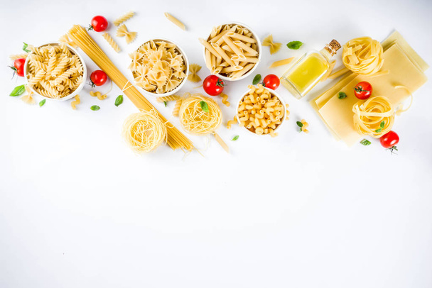 Italian food concept, various raw pasta assortment - spaghetti, lasagna, fusilli, tagliatelle, penne, tortellini, ravioli, with tomatoes and basil leaves white background copy space top view - Photo, Image