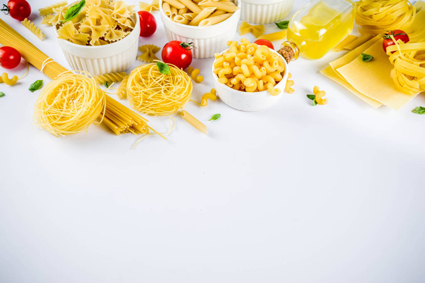 Italian food concept, various raw pasta assortment - spaghetti, lasagna, fusilli, tagliatelle, penne, tortellini, ravioli, with tomatoes and basil leaves white background copy space top view - Photo, Image