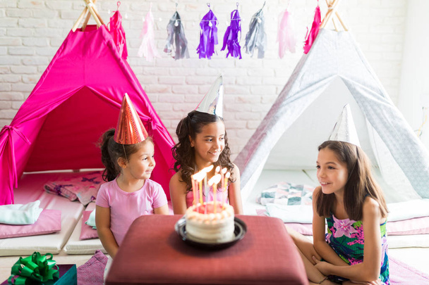 Leuke meisjes dragen feestmutsen met verjaardagstaart op tafel tegen tipi tenten thuis - Foto, afbeelding