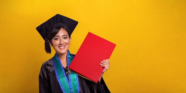 Young attractive Asian female student on her graduation day, holding a red colored certificate with happy face, over bright yellow studio background - Photo, Image