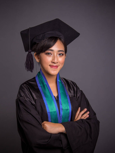 卒業キャップ アンド ガウンで若い魅力的なアジア女性独身の肖像画、色背景のグレーが自信を持って、交差の腕のポーズ - 写真・画像