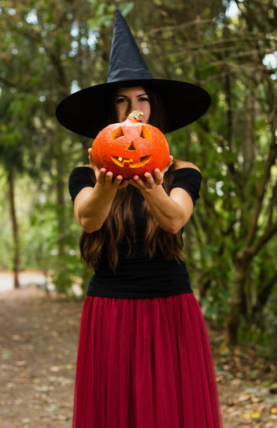 Halloween vakantie achtergrond. Halloween heks met een halloween pompoen jack o lantern decor met grappige gezicht in een donker bos. Mooie jonge vrouw in heksen hoed en houden van pompoen kostuum. - Foto, afbeelding