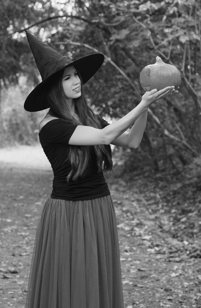Tło wakacje Halloween. Witch Halloween halloween dynia jack o lantern urządzone z śmieszną twarz w ciemnym lesie. Piękna młoda kobieta w kapelusz czarownic i trzymając dynia kostium. Czarno-białe zdjęcie. - Zdjęcie, obraz