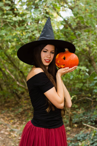 Halloween holiday background. Halloween Witch with a halloween pumpkin jack o lantern decor with funny face in a dark forest. Beautiful young woman in witches hat and costume holding pumpkin. - Photo, Image