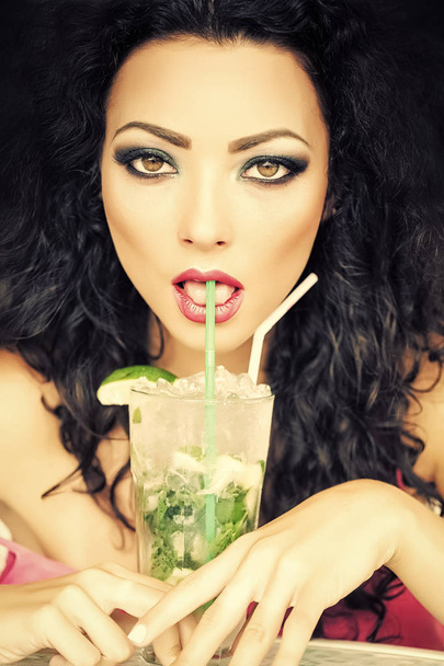 Portrait of cute young brunette woman with curly hair and bright makeup drinking alcoholic mojito cocktail from mint soda light rum ice cubes and lime with straw, vertical picture - Foto, Imagen