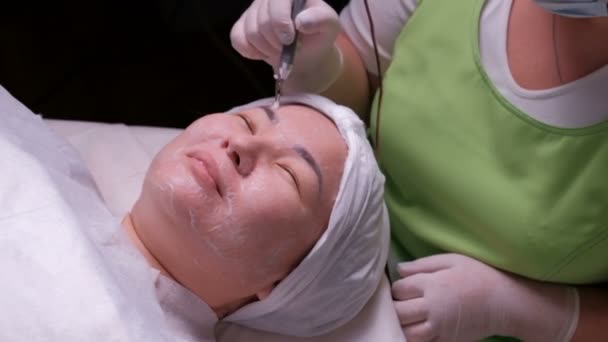Galvanic cleaning of the skin. Asian woman on the procedure of disincrustation. The work of a beautician with an electrical apparatus in hands. Oriental girl at the reception in a medical beauty salon - Footage, Video