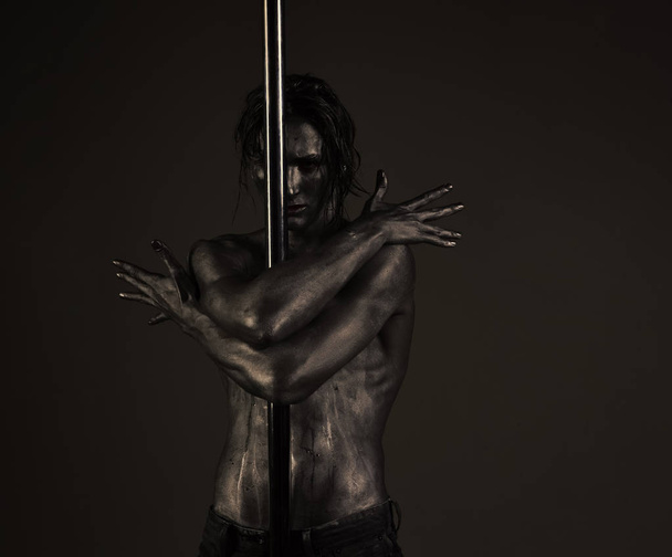 Macho with naked chest, athlete, sportsman performing gentle pole dancing moves. Man with nude torso covered with silver paint, black background. Guy hugs metallic pole. Passionate dancer concept. - Фото, зображення