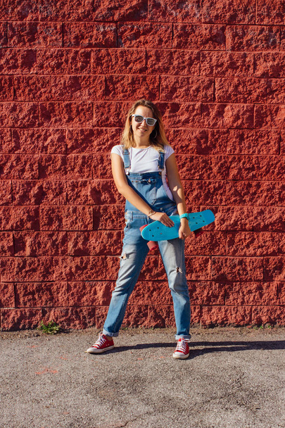 Portrait of a smiling woman dressed in overalls and sunglasses standing with her skateboard next to the red wall. - Фото, изображение