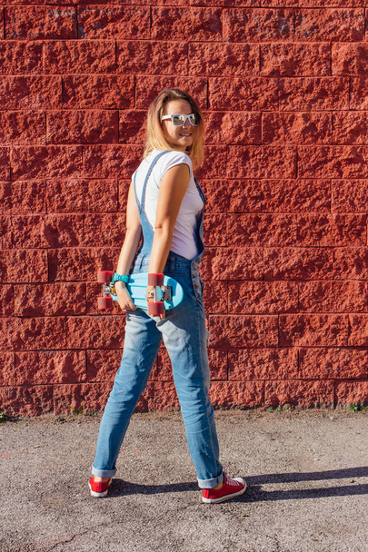 Portrait of a smiling woman dressed in overalls and sunglasses standing with her skateboard next to the red wall. - Foto, Bild