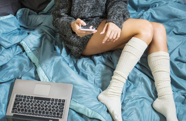 Młoda dziewczyna z sexy nogi w ciepły sweter, leżąc na łóżku z smartphone i laptopa. Praca w domu. Widok z góry - Zdjęcie, obraz