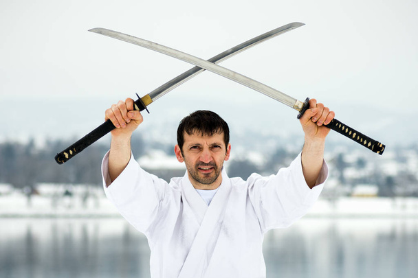 Combatiente en kimono blanco sostiene dos espadas japonesas al aire libre en invierno
. - Foto, imagen