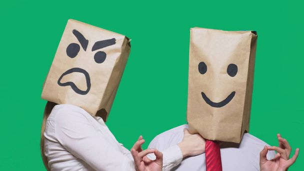 The concept of emotions and gestures. Two people in paper bags on the head with painted smileys. Aggressive smiley swears. The second smiles at him. - Photo, Image