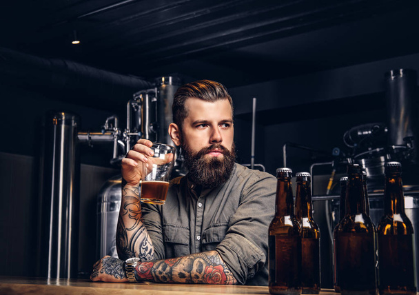 Tattooed hipster male with stylish beard and hair drinking beer sitting at the bar counter in the indie brewery. - Photo, Image