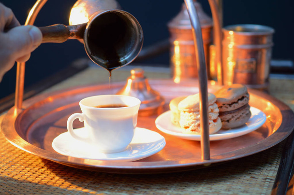 Kaffee aus einer Kanne in eine weiße Tasse neben der Kupferkaffeemaschine und Makronen gießen - Foto, Bild