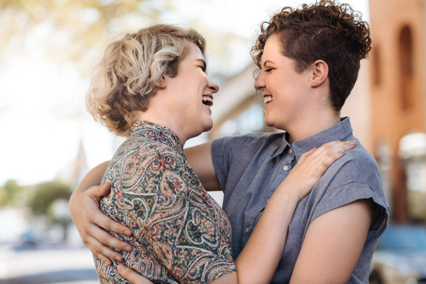 Laughing young lesbian couple standing arm in arm together on a city street listening to music on earphones - 写真・画像