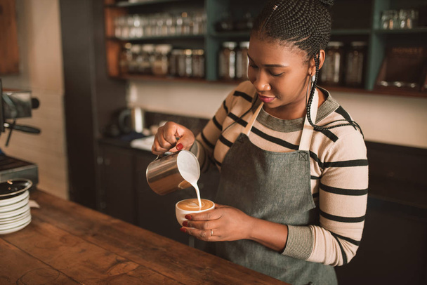 Smiling young African barista standing behind the counter of her trendy cafe pouring frothed milk into a cup of cappuccino  - Photo, Image