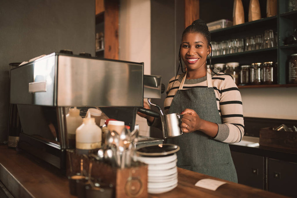 Portrait of a smiling young African barista frothing milk for a cappuccino behind the counter of her trendy cafe - 写真・画像