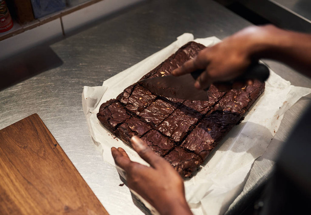 Closeup of a baker cutting delicious brownies into portions while working at a counter in a commercial bakery  - 写真・画像