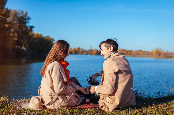 Νεαρό ζευγάρι στην αγάπη ψύξη από φθινόπωρο στη λίμνη. Ευτυχισμένος άνθρωπος και γυναίκα απολαμβάνοντας τσάι σε εξωτερικούς χώρους. Ανθρώπους να πίνουν καφέ σε πικ-νικ - Φωτογραφία, εικόνα