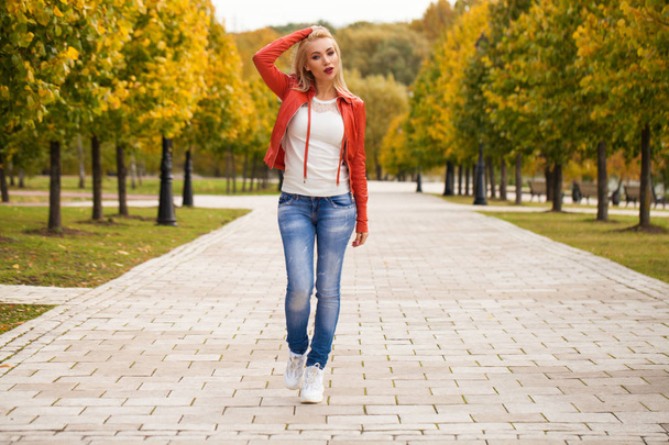 Portrait of happy smiling beautiful young blonde woman in blue jeans and red jacket, autumn street outdoors - Fotoğraf, Görsel