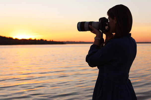 Young female photographer taking photo of riverside sunset with professional camera outdoors - Foto, afbeelding