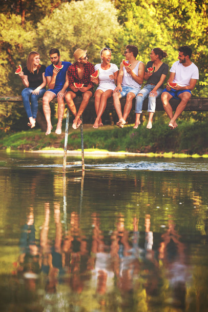 groupe de jeunes amis profitant de pastèque tout en étant assis sur le pont en bois sur la rivière dans une belle nature
 - Photo, image