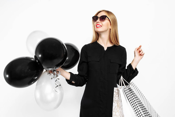 happy woman in sunglasses in black dress and with red lips holding bags and black balls, satisfied girls after shopping, Black Friday on white background - Foto, Bild