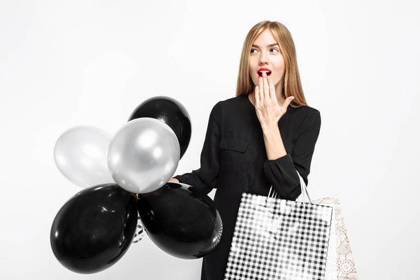 Elegant shocked girl, in black dress, with shopping bags and black balls in her hands isolated on white background . Black Friday, shopping, discounts - Zdjęcie, obraz
