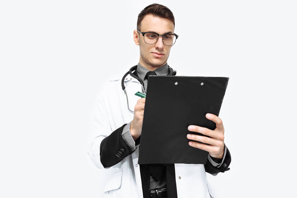 a young Intern with glasses, a white robe and a stethoscope around his neck, takes notes in the medical records, and smiles on a white background - Zdjęcie, obraz