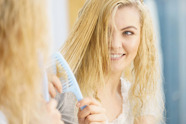 Young blonde woman brushing her long wet hairdo using hair brush. Haircare and hairstyling concept. - Fotó, kép