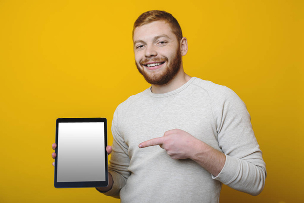 Gyömbér szakállal és mosolyogva néz a kamera állva, fényes, sárga háttér előtt, és bizonyítja a modern tabletta-val üres képernyő, csinos, fiatal férfi - Fotó, kép