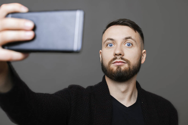 スタイリッシュなコスチュームでスマート フォンを保持している灰色の背景に selfie のポーズをしながらカメラ目線でハンサムな男性 - 写真・画像