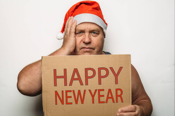 Portrait of funny senior man in red Santa Claus Christmas hat holds cardboard with inscription Happy New Year in hands, holds his hand on head and looks at camera, Humor festive and celebrate concept - Photo, Image