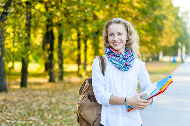 Happy charming young woman with backpack standing and holding notebooks in park - Photo, Image