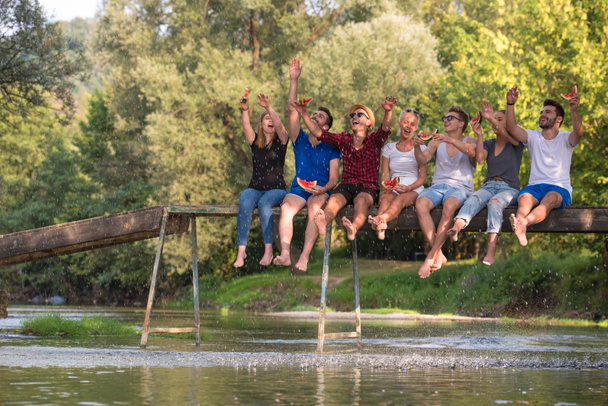 groep van jonge vrienden genieten van watermeloen zittend op de houten brug over de rivier in prachtige natuur - Foto, afbeelding