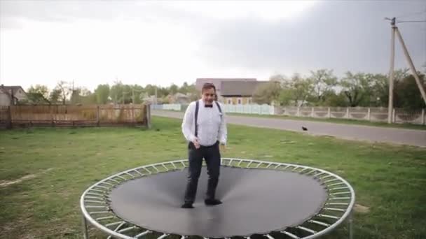 Een jonge man in zwarte broek met bretels en een wit overhemd met een bowtie springt op een trampoline - Video